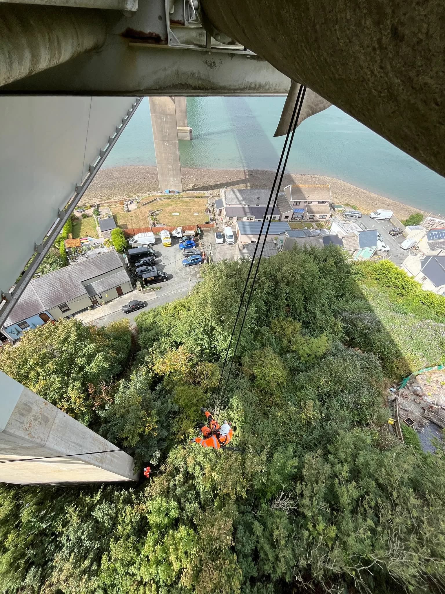 Looking down from beneath the bridge at a technician descending over the wooded bank and the houses below