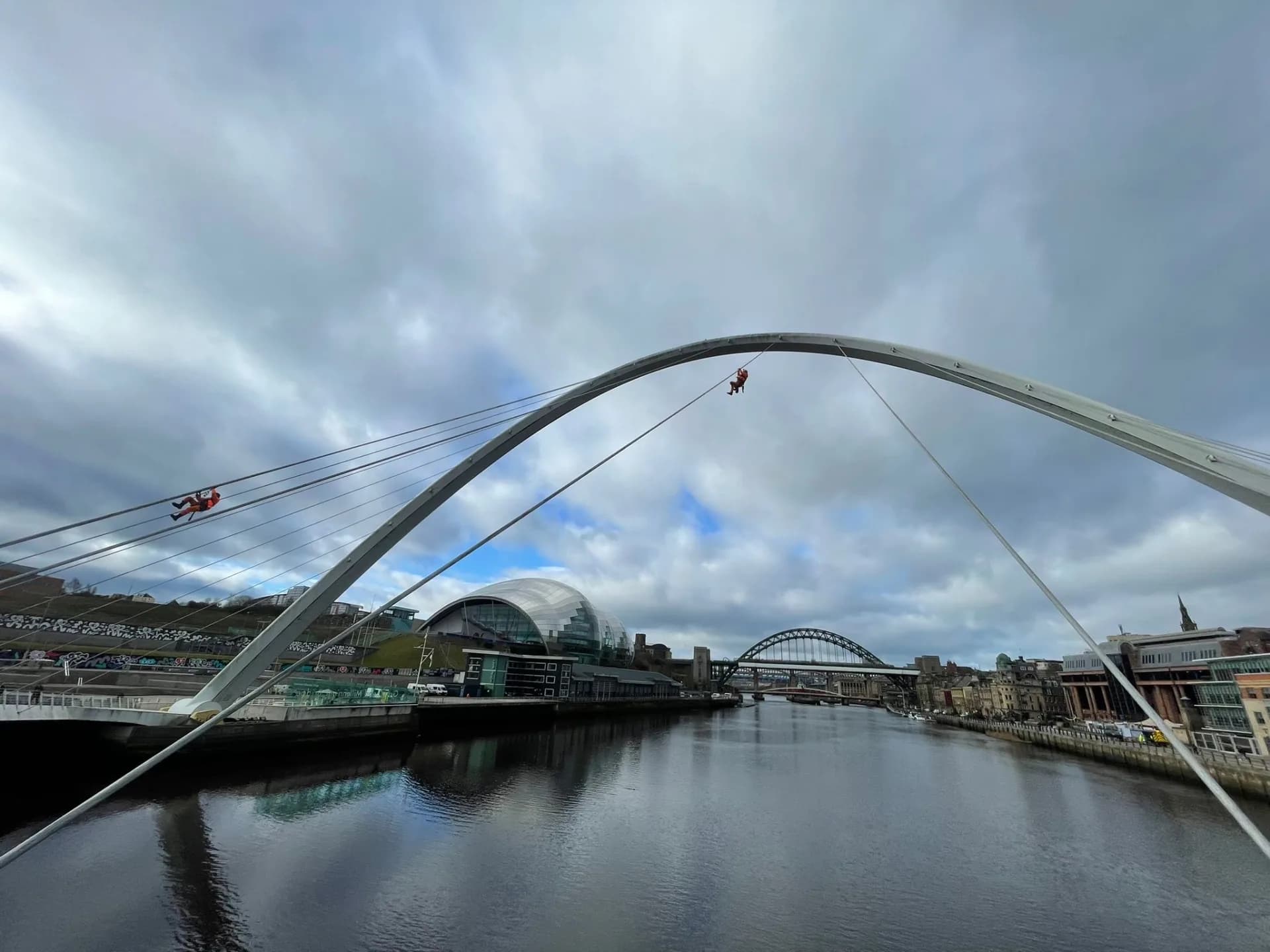 Two rope access technicians working on the arch and cables of the Gateshead Millennium Bridge, with the Tyne Bridge beyond