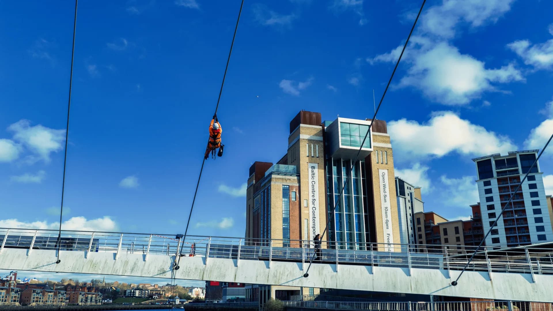 Technician on a cable high above the bridge deck, against a blue sky and the Baltic Centre