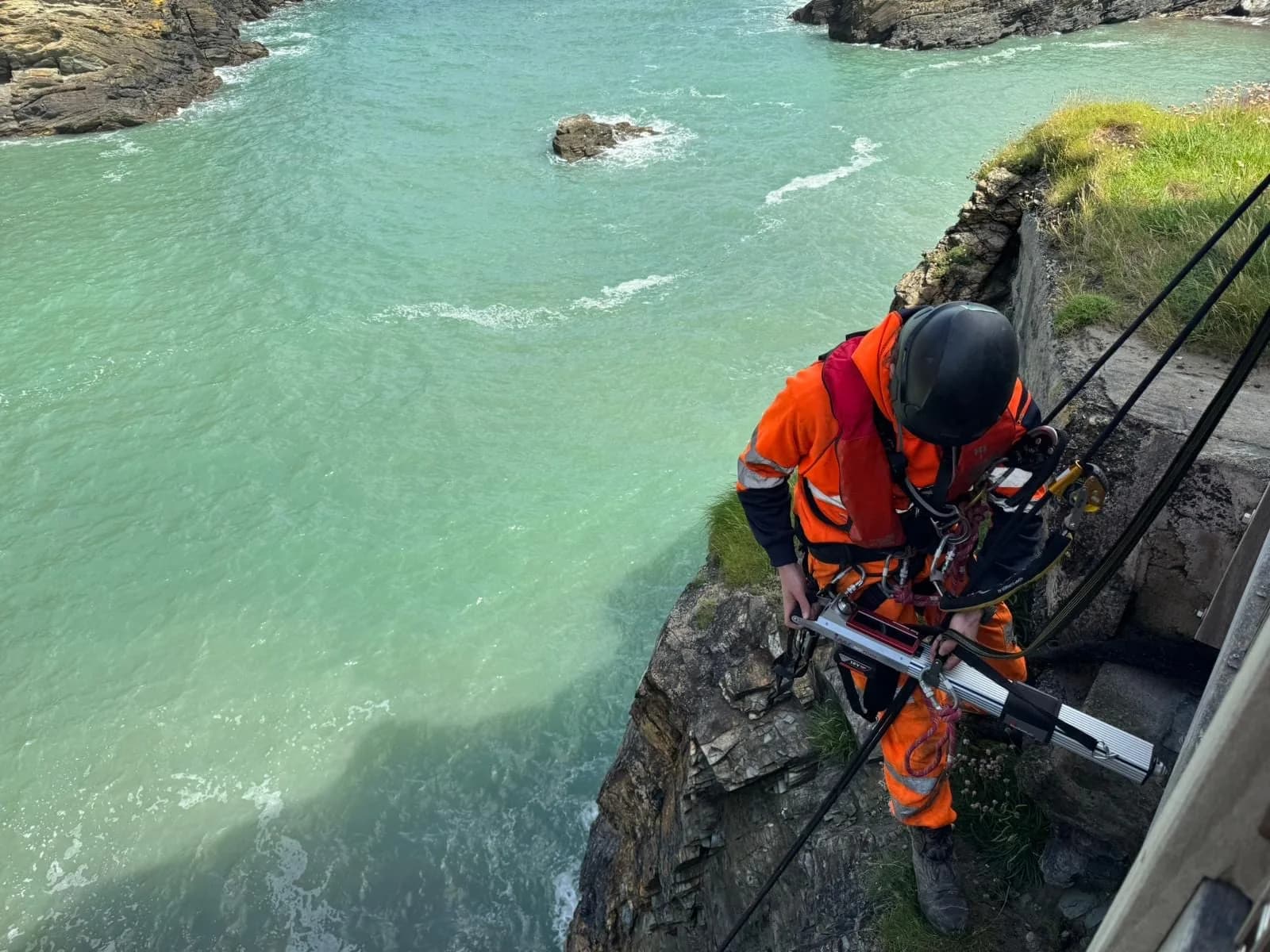Rope access technician operating test equipment on a cliff edge above the sea